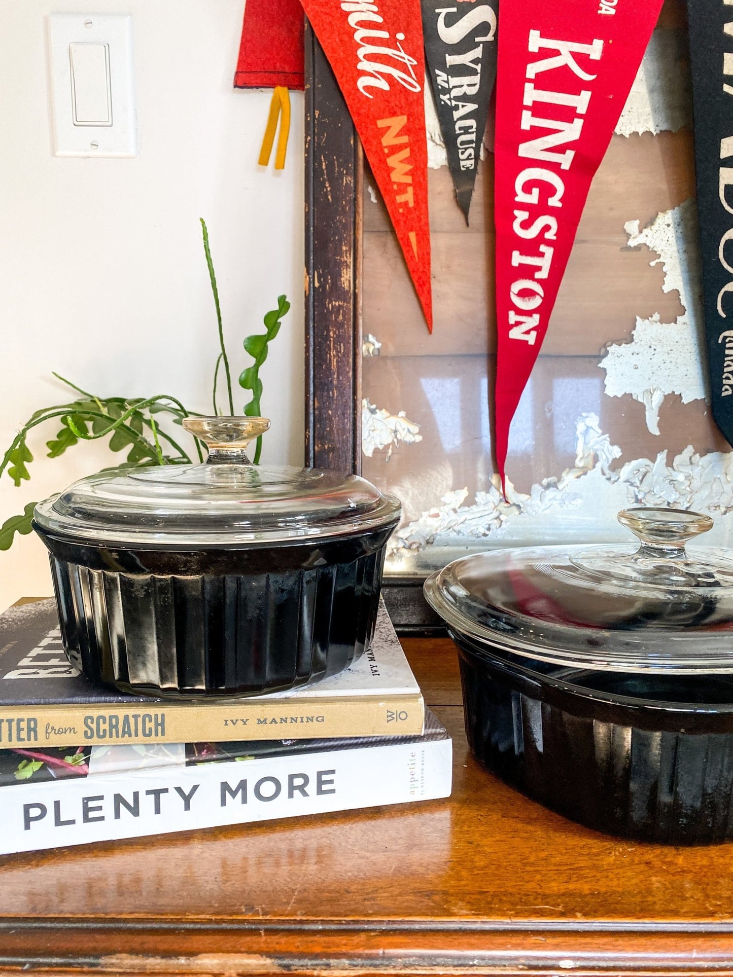 There are two black dishes in focus with the smaller round on on top of two cookbooks, Better from Scratch and Plenty More. The other black casserole dish is off to the right with the lid partly off. The background features vintage pennant flags for Fort Smith, NWT, Syracuse, NY and Kingston.