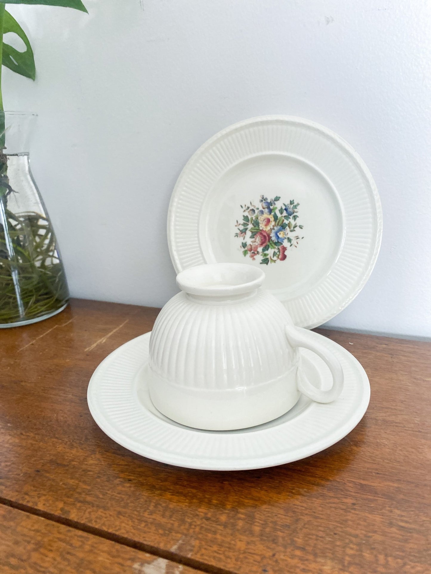 A teacup sits on upside down on top of a saucer, it shows the exterior of the cup on the side. The lunch plate is at the back standing upright to show the floral design. There is a monstera plant in the back left.